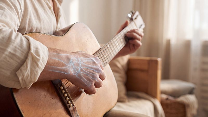 Hands playing an acoustic guitar, with the bones of the hand and wrist showing softly through the skin in blue.