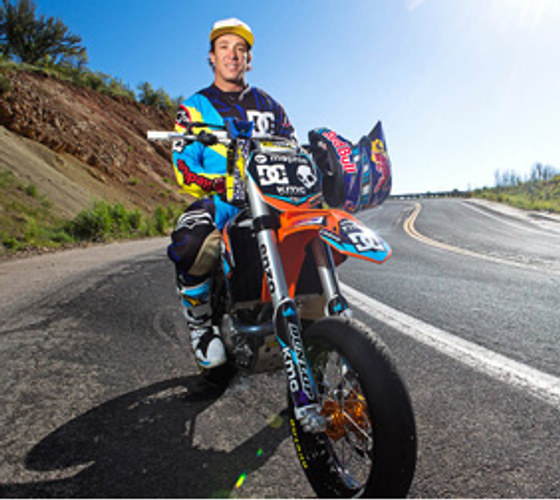 Robbie Maddison in race gear, sitting astride his motocross bike on a hillside road, Red Bull and DC branding on the bike.