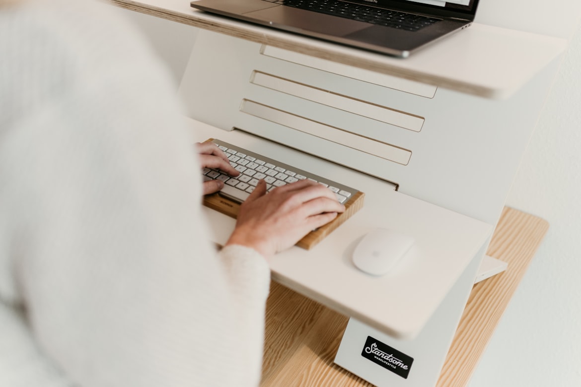 A pair of hands at a keyboard on a white desk, seen from above, with a laptop on a stand behind them.
