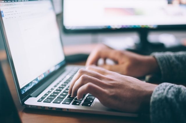 Hands typing on a laptop keyboard, with a second screen out of focus behind them.